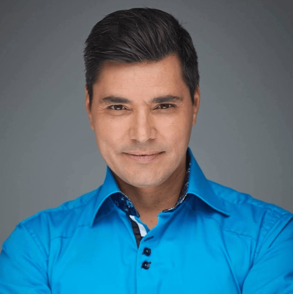Hombre sonriendo con camisa azul, fondo gris.