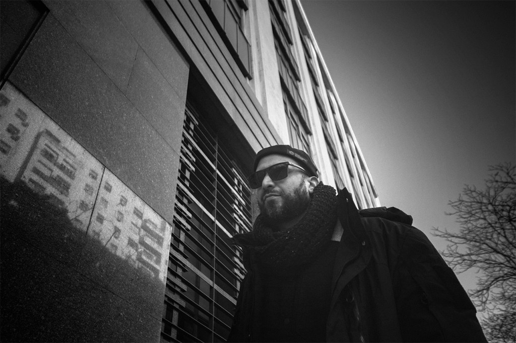 Hombre con barba y gafas de sol, caminando junto a un edificio en tonos grises, con reflejos en la pared.