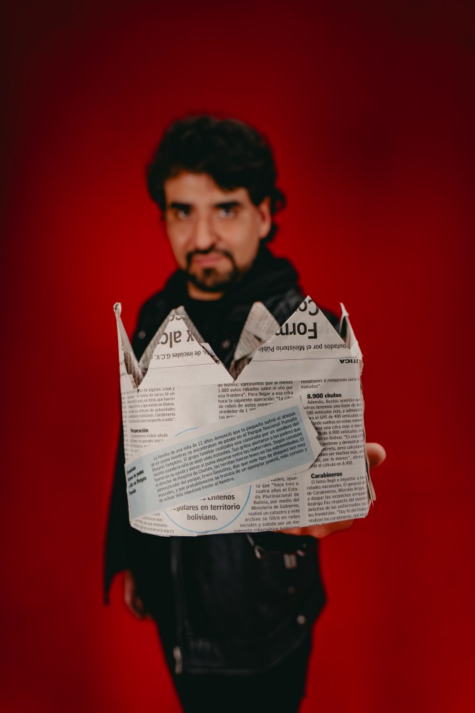 Un hombre sostiene una corona de papel elaborada con periódico frente a un fondo rojo.
