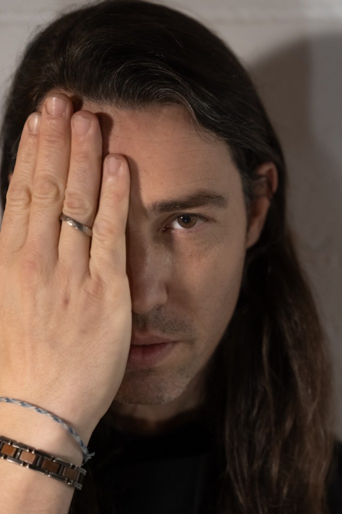 Retrato de un hombre con cabello largo, cubriendo parcialmente su rostro con una mano, mirando directamente a la cámara.