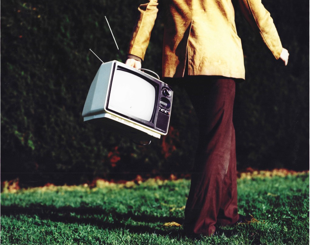 Persona caminando por un césped sosteniendo un televisor antiguo.