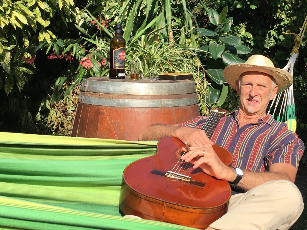 Hombre sentado en una hamaca, tocando una guitarra, con un barril de madera y una botella de ron en el fondo, rodeado de vegetación.