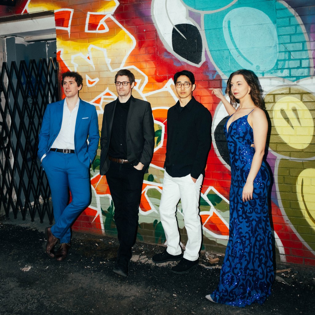 Grupo de cuatro personas posando frente a un mural colorido, con un hombre vestido con un traje azul brillante, otro en un traje negro, un tercer hombre con camisa negra y pantalones claros, y una mujer con un vestido azul brillante.