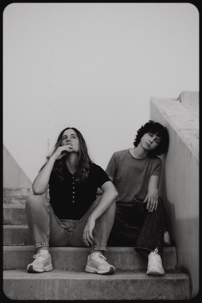 Dos personas sentadas en escalones, una sosteniendo su mentón y mirando pensativa, y la otra con expresión relajada. La imagen es en blanco y negro.