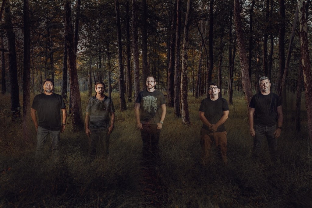 Cinco hombres de pie en un bosque, rodeados de árboles y hierba alta, con luz tenue que ilumina la escena.