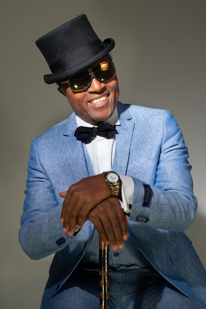 Hombre sonriente con un sombrero de copa y gafas de sol, vestido con un traje azul claro y pajarita negra, posando con un bastón en un fondo neutro.