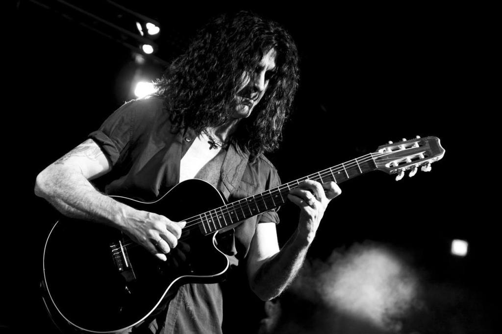 Músico tocando una guitarra negra en un escenario, iluminado con luces brillantes.